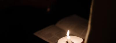 Taper candle casting glow on person and paper program at a prayer vigil.